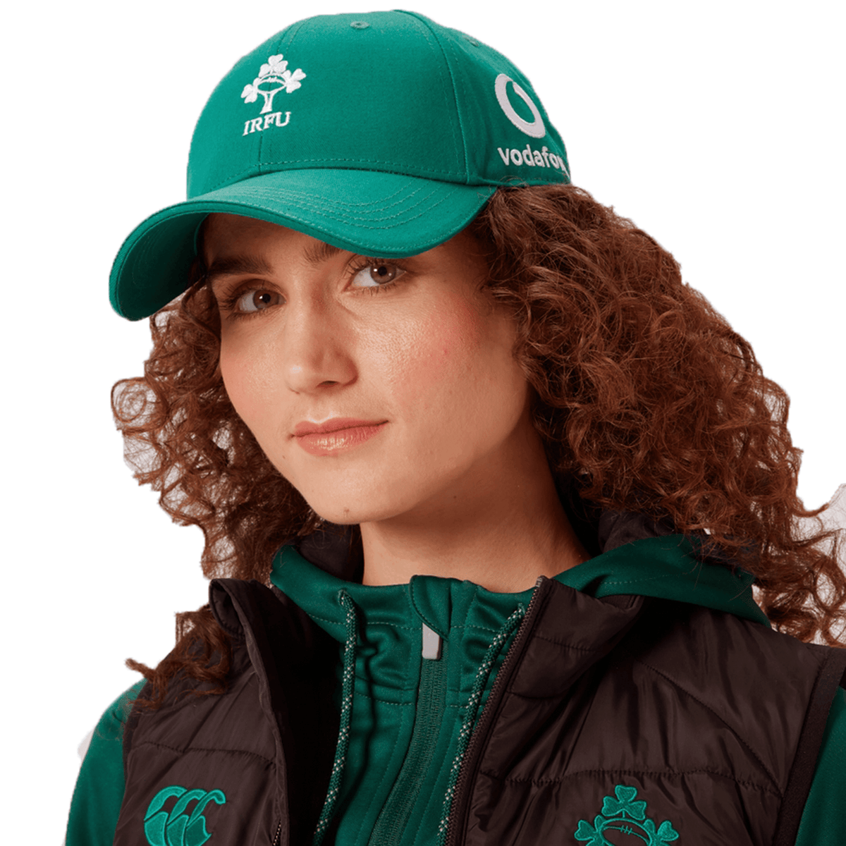 A person with curly hair wears the Ireland 25/26 Drill Cap by Canterbury in green, plus a matching jacket and black vest featuring Vodafone and IRFU logos.