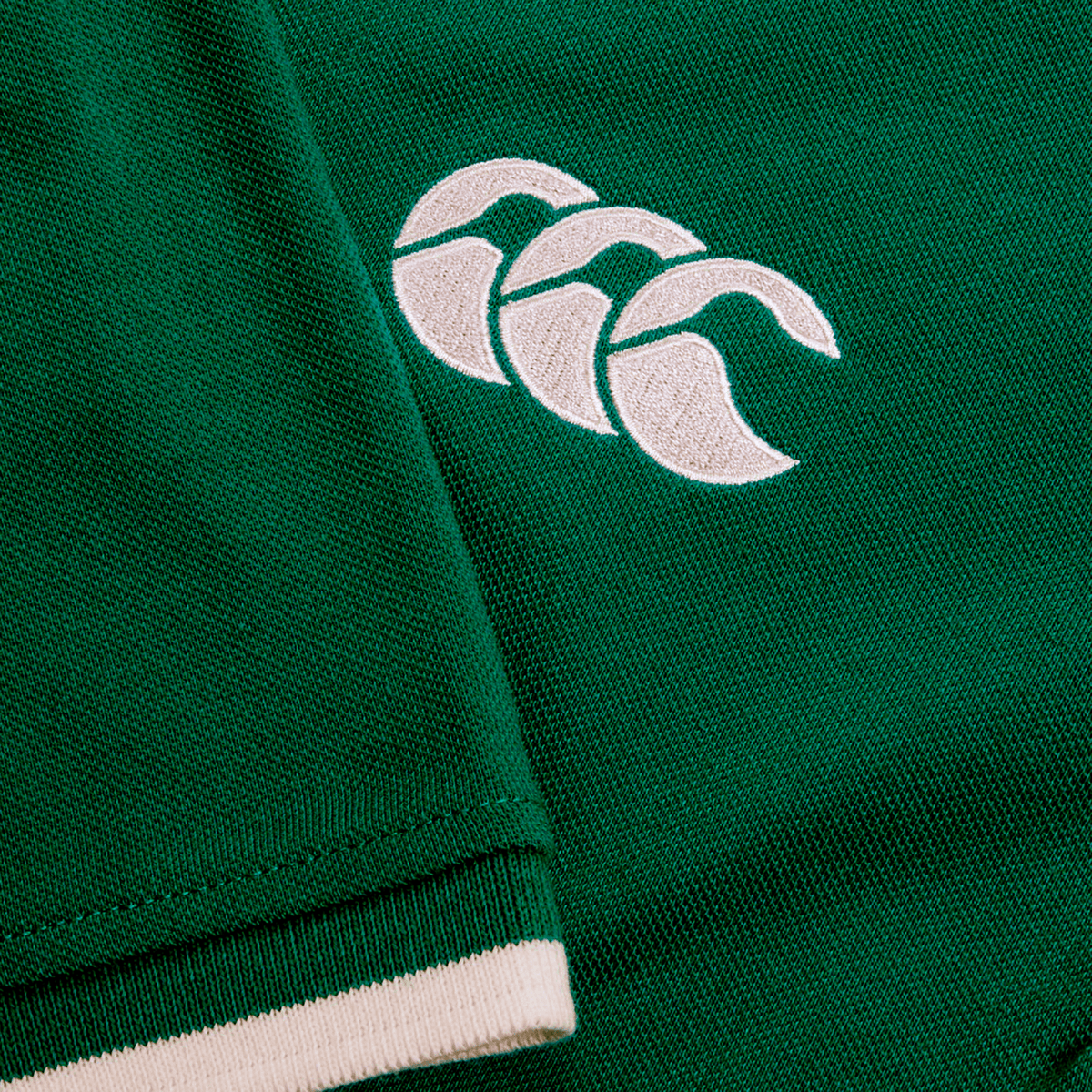 Green fabric from the Ireland 25/26 Team Polo by Canterbury with a white embroidered logo of three overlapping curved shapes. Visible stitching appears near a folded seam highlighted by a white trim.