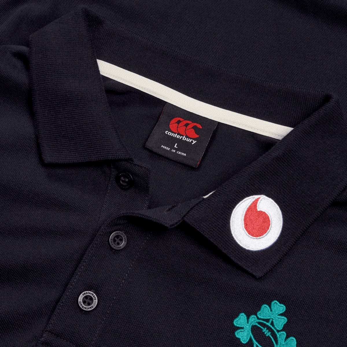 Close-up of the black Ireland 25/26 Team Polo by Canterbury, featuring buttons, an embroidered Vodafone logo on the collar, and a green shamrock emblem on the chest.