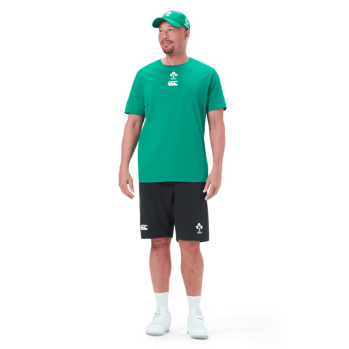 A man wearing the Ireland Team Cotton T-Shirt 24 by Canterbury and black shorts stands, adorned with a green cap that has an Irish green logo, and white sneakers.