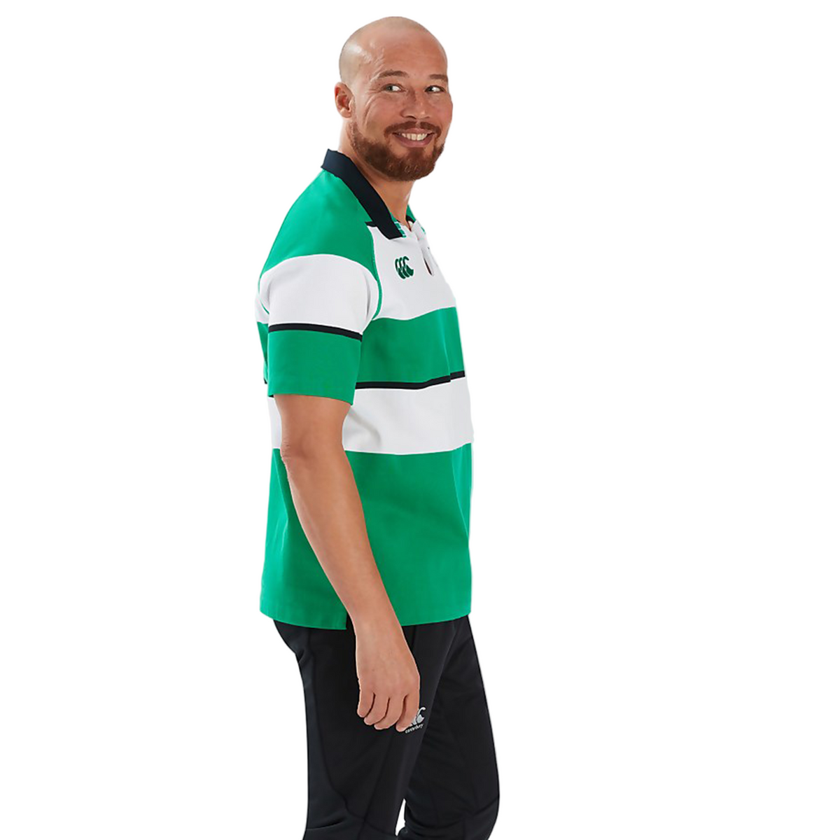 A man with a shaved head and beard, wearing the Canterbury Ireland 25/26 Heritage Hooped Jersey in green and white stripes and black pants, stands smiling and facing slightly to the side.