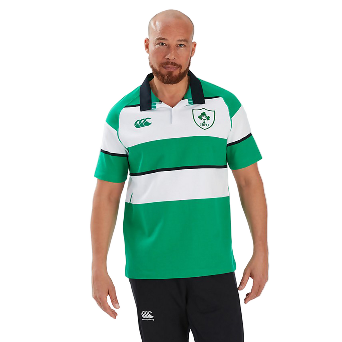 A man with a shaved head and beard wears the Canterbury Ireland 25/26 Heritage Hooped Jersey—green and white stripes with the Ireland crest—and black athletic pants, standing against a plain light grey background.