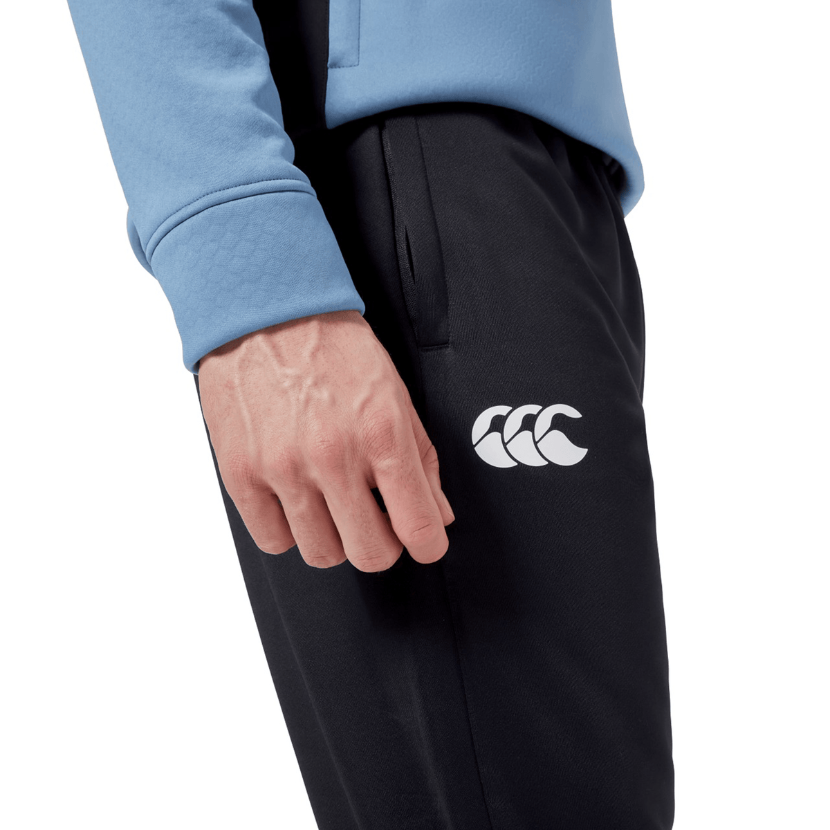 Close-up of a person wearing a light blue jacket and Ireland 24 Poly Knit Pant by Canterbury, which features VapoDri technology. The black pants have a white Canterbury logo depicting three overlapping shapes on the side.