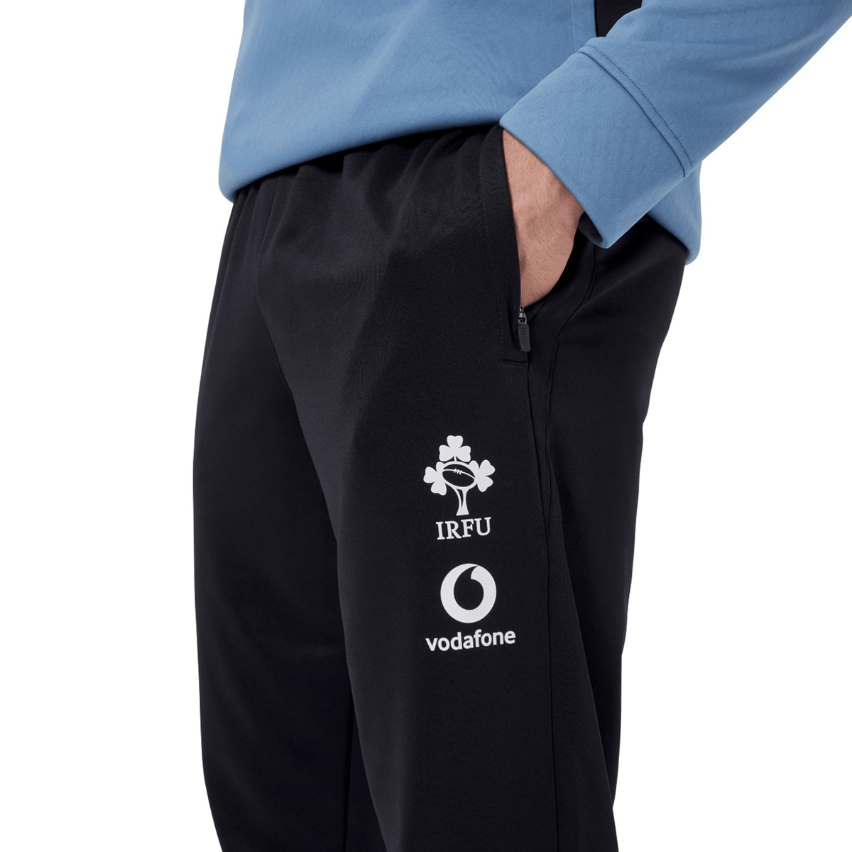 An Irish rugby fan with their hand in the pocket of Canterbury&#39;s Ireland 24 Poly Knit Pant, showcasing the IRFU and Vodafone logos, is wearing a long-sleeve blue top equipped with VapoDri technology.