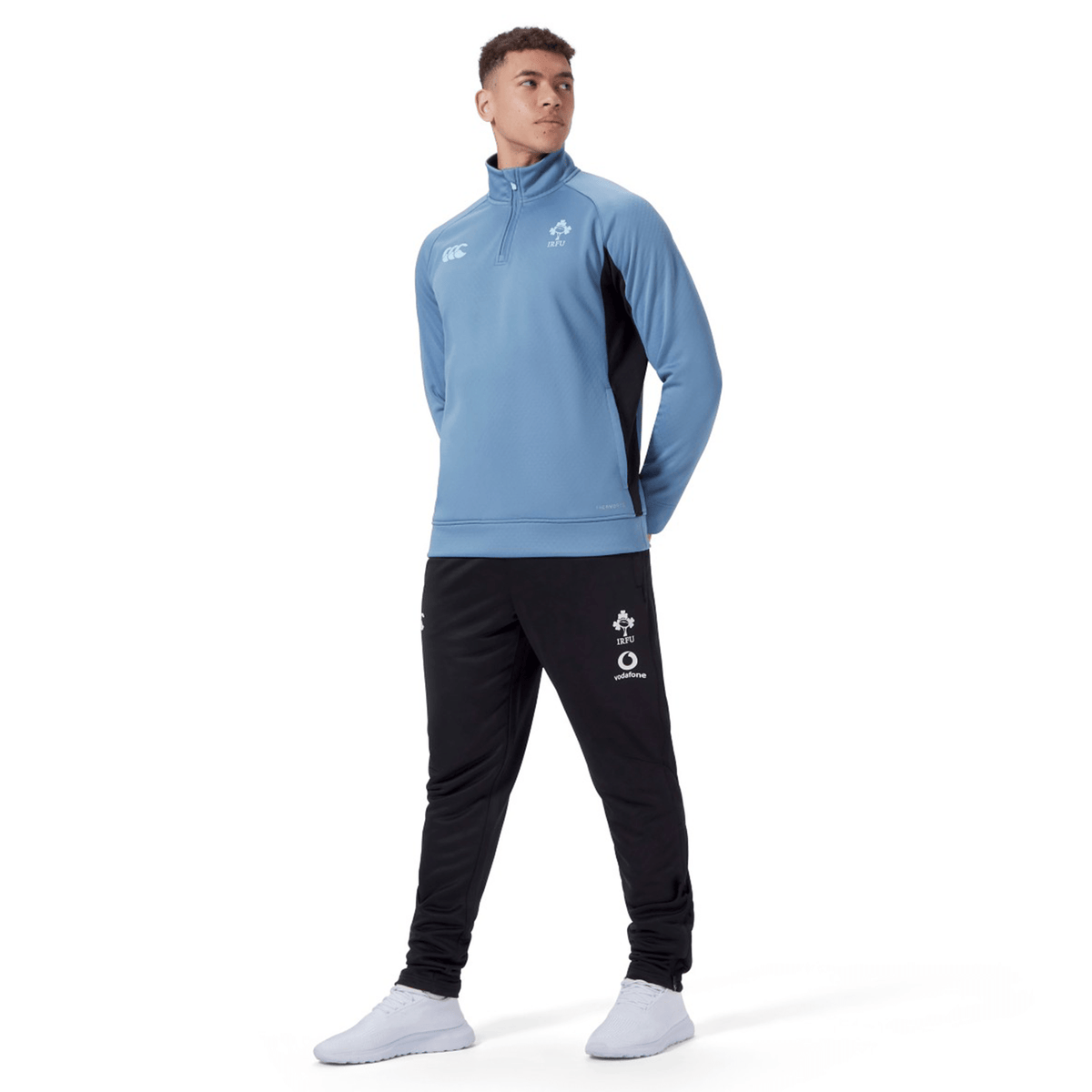 A person dressed in a blue quarter-zip tracksuit top and the Ireland 24 Poly Knit Pant by Canterbury, which features VapoDri technology, is standing against a white background. They are facing slightly to the right with their hands behind their back.