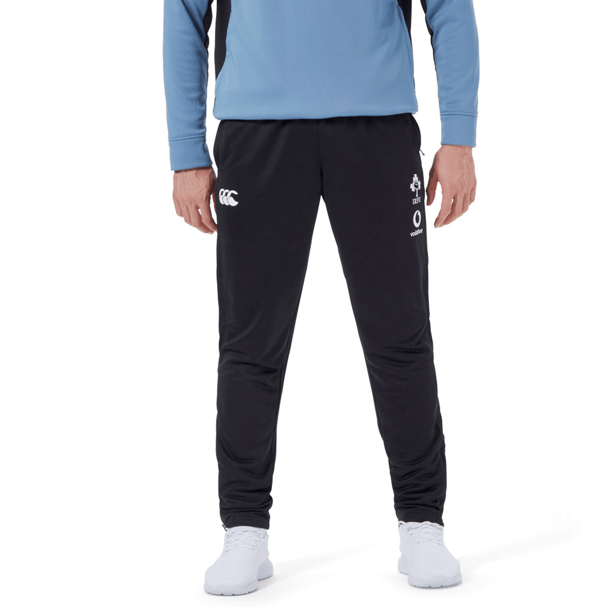 A person wearing black Ireland 24 Poly Knit Pant by Canterbury with white logos on the thighs, a light blue top, and white shoes.