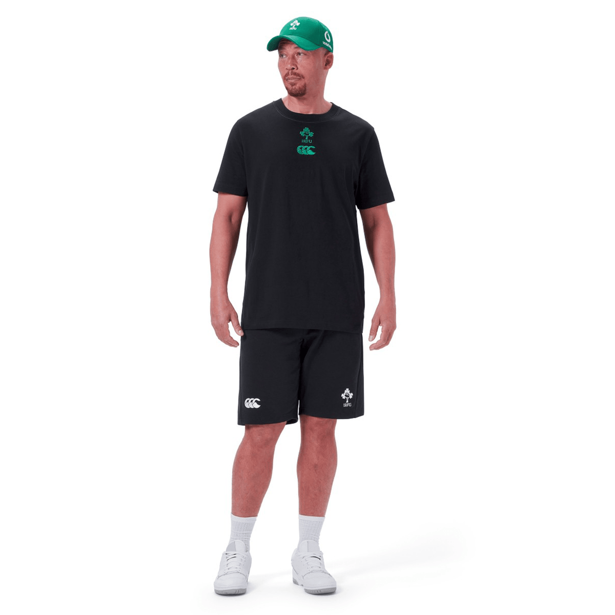 A man wearing a black Ireland Team Cotton T-Shirt 24 by Canterbury, black shorts, white socks, white shoes, and a green baseball cap with green logos stands against a white background.