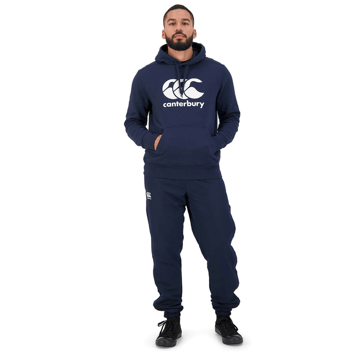 A person stands wearing a navy blue Canterbury Anchor Hoodie and matching sweatpants, hands in pockets, and black shoes. The fleece-lined hoodie from Canterbury adds an extra layer of warmth, perfect for cool weather.