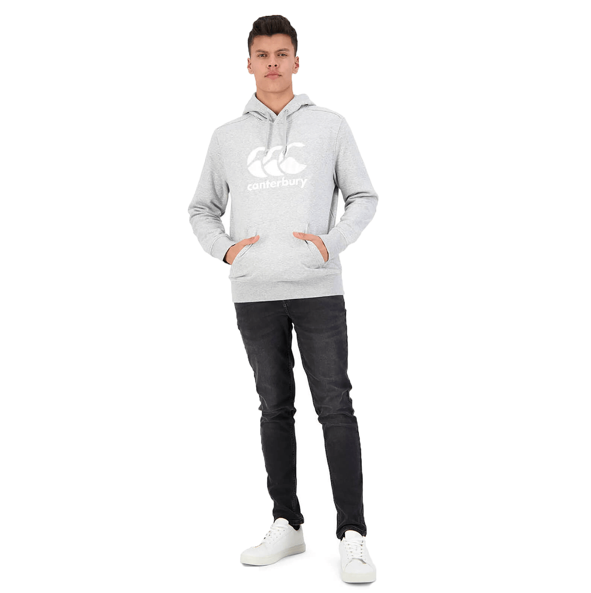 A man standing wearing a light gray Canterbury Anchor Hoodie with a white logo, black jeans, and white sneakers.