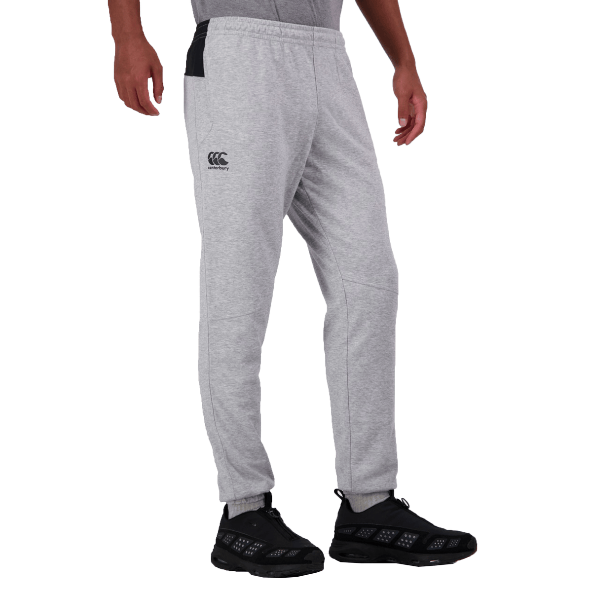 The image depicts a person waist down, wearing Canterbury's light gray Tempo Vapodri Hybrid Cuffed Pant with tapered legs and an elasticated waistband featuring a logo on the left thigh, paired with sleek black sneakers against a crisp white background.