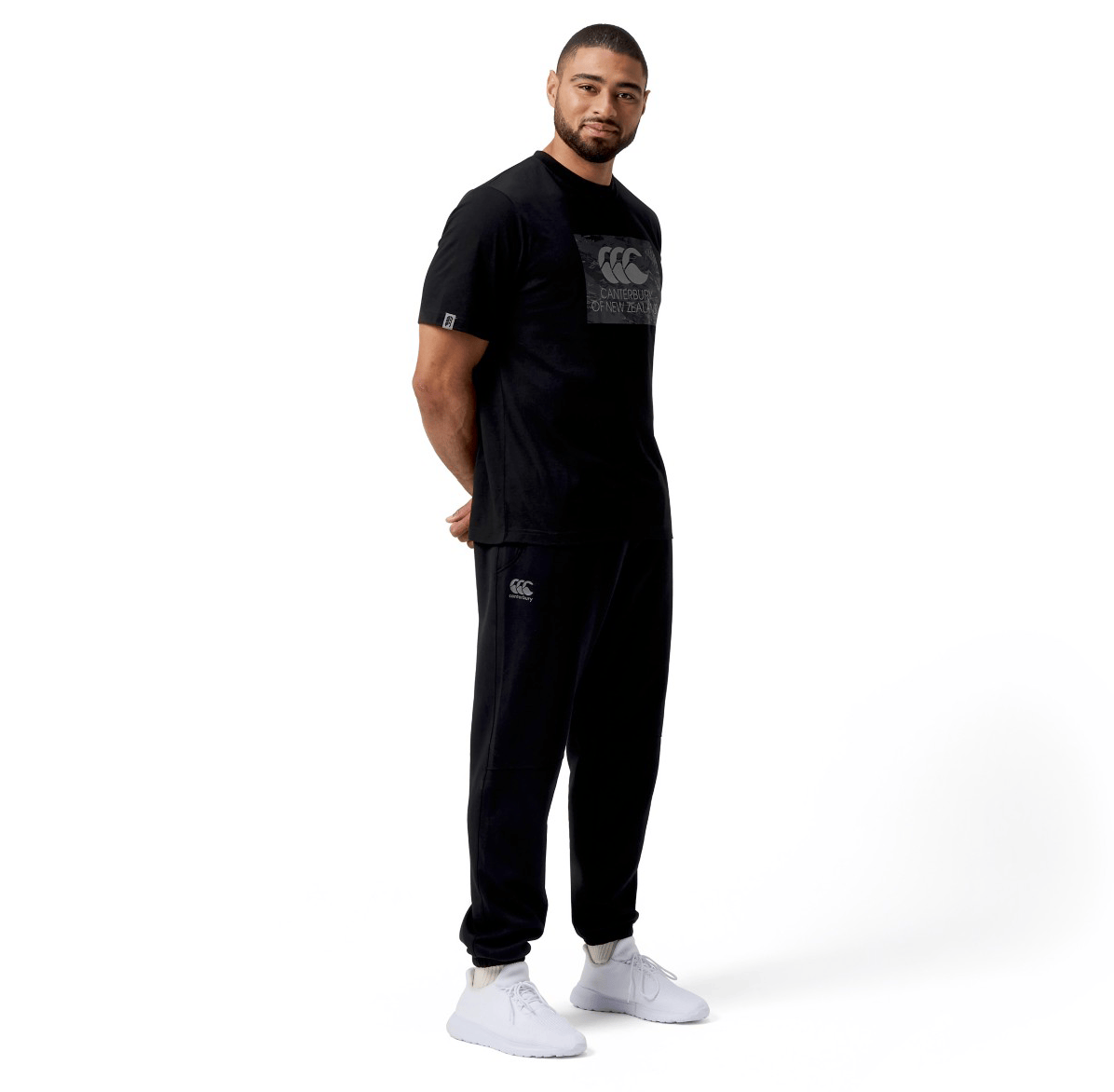 A man stands against a white background wearing a black t-shirt, Canterbury Tempo Vapodri Hybrid Cuffed Pant with a tapered leg, and white sneakers.