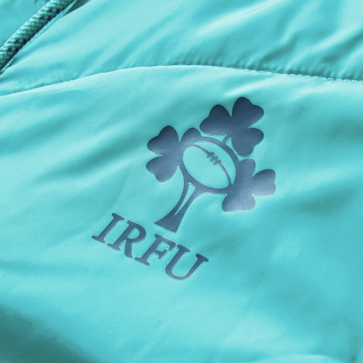 Close-up of a teal Ireland 26 Hybrid Padded Jacket by Canterbury, featuring the IRFU logo with a rugby ball and shamrock design printed above the letters “IRFU.”.