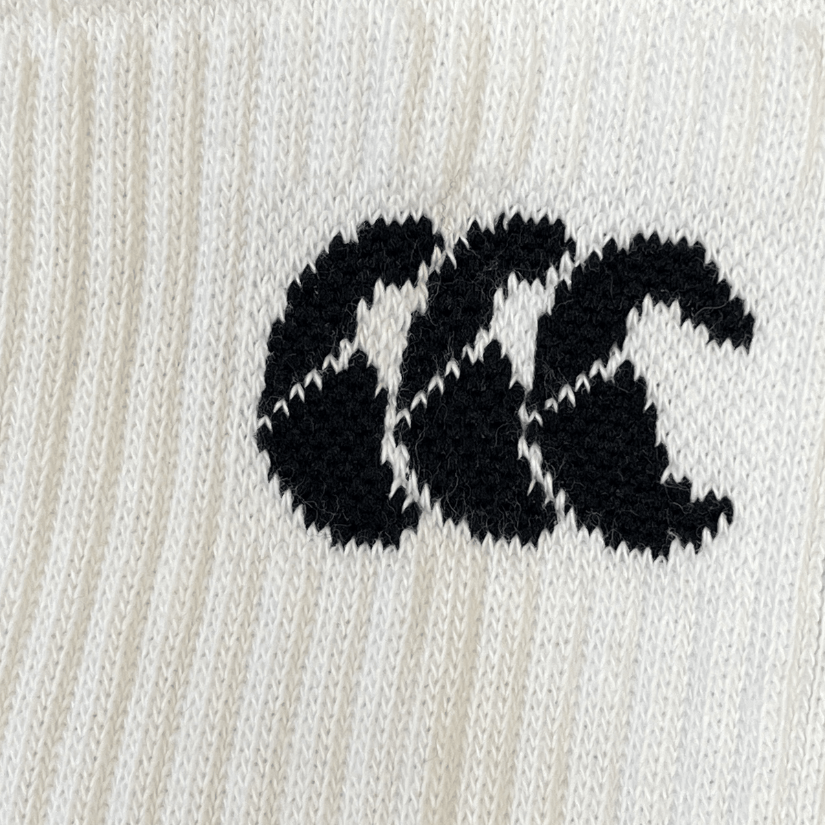 Close-up of a white knitted fabric Canterbury Mid Calf Grip Sock featuring a black embroidered logo with three overlapping stylized C's and enhanced arch support using Premgripp technology.