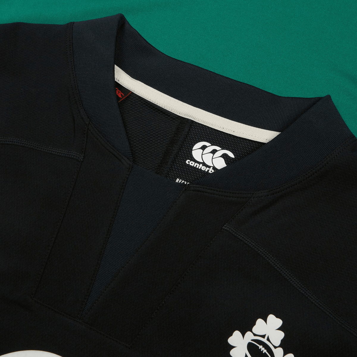 Close-up of the Ireland 25/26 Tech Drill Top by Canterbury, a black rugby top with the Canterbury logo and white shamrock emblem on the chest, shown against a green background.