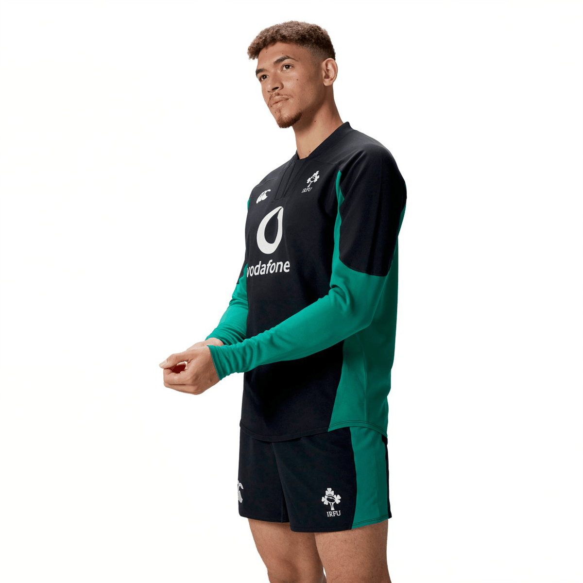 A man wears the Ireland 25/26 Tech Drill Top by Canterbury in black and green, standing against a white background and looking to the side with his hands together.
