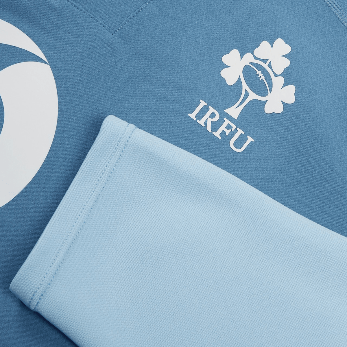 Close-up of the blue Ireland 25/26 Tech Drill Top by Canterbury, showcasing the IRFU logo with rugby ball and shamrock design, plus part of a white circular emblem on the left.