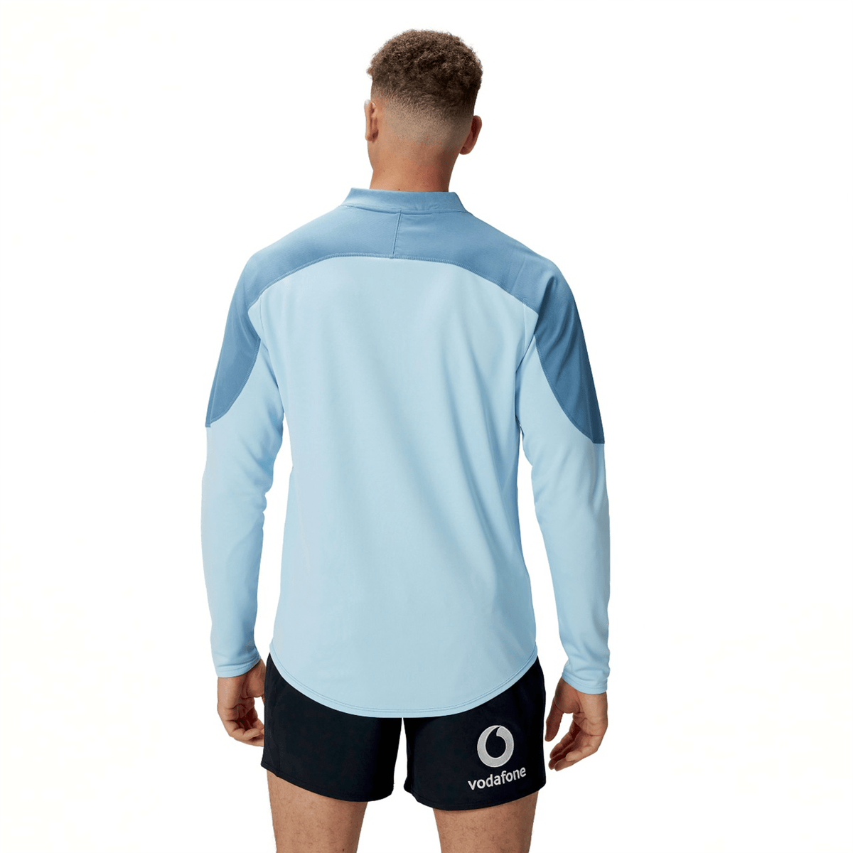 A person stands facing away, wearing the Ireland 25/26 Tech Drill Top by Canterbury—a light blue rugby top with darker blue shoulder panels—and black shorts with a Vodafone logo.