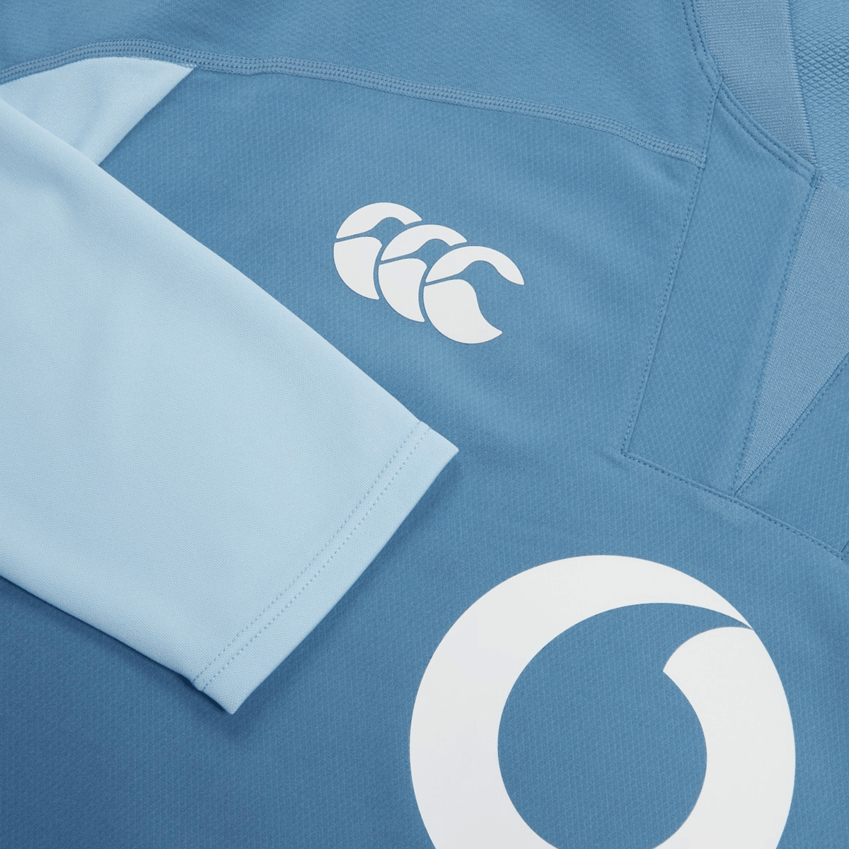 A close-up of the blue Canterbury Ireland 25/26 Tech Drill Top highlights the iconic logo and a partial white circular front design, showcasing the detail of this official Canterbury rugby top.