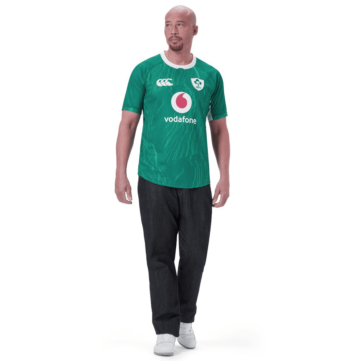 A bald man with a short beard is wearing the green Ireland 24 Home Pro Jersey by Canterbury, black pants, and white sneakers. He is standing and looking to his left.
