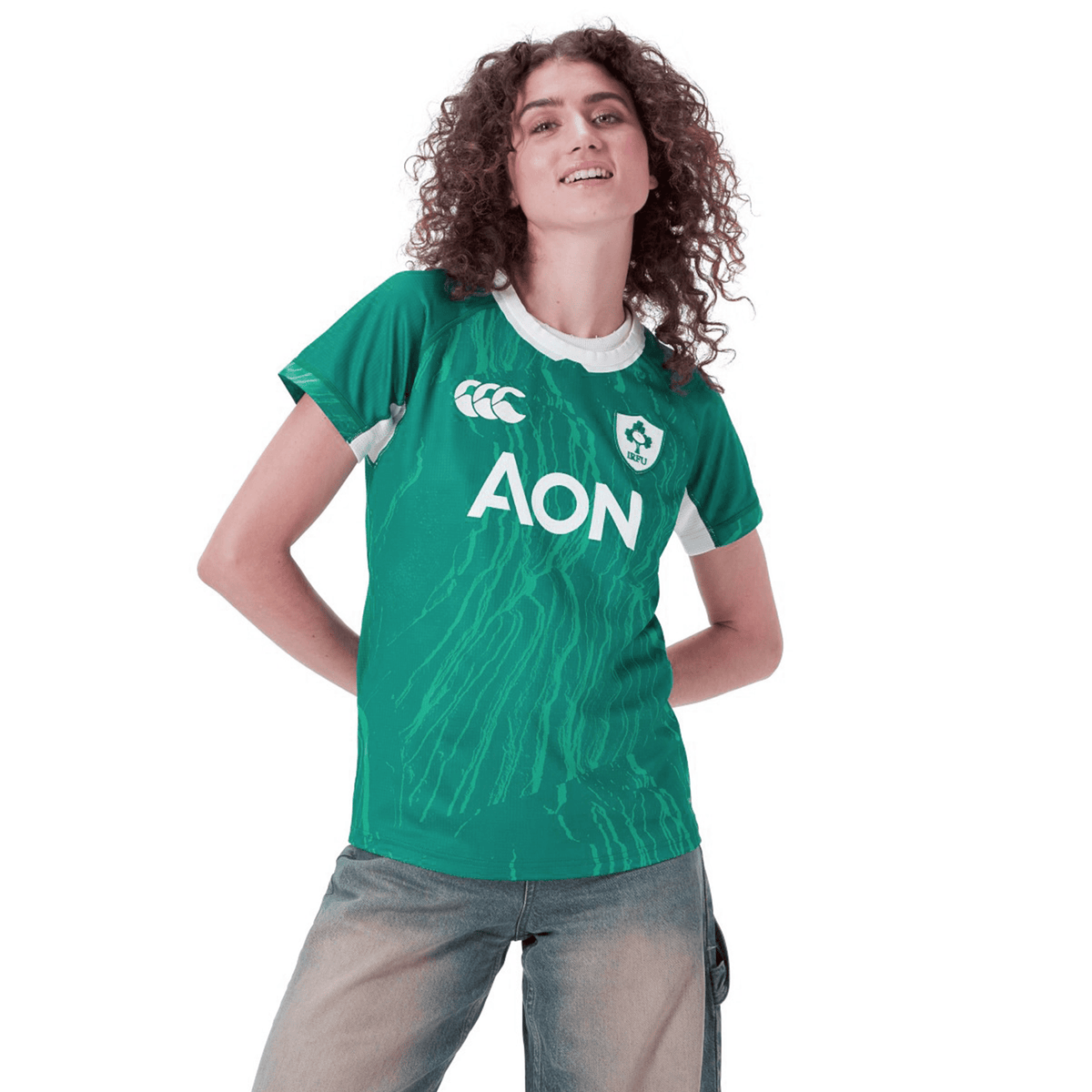 A person is wearing a green Women&#39;s Ireland WIRFU Home Pro Jersey by Canterbury, featuring VapoDri technology and the &quot;AON&quot; logo. They pair it with light jeans against a white background, proudly displaying the official IRFU crest.