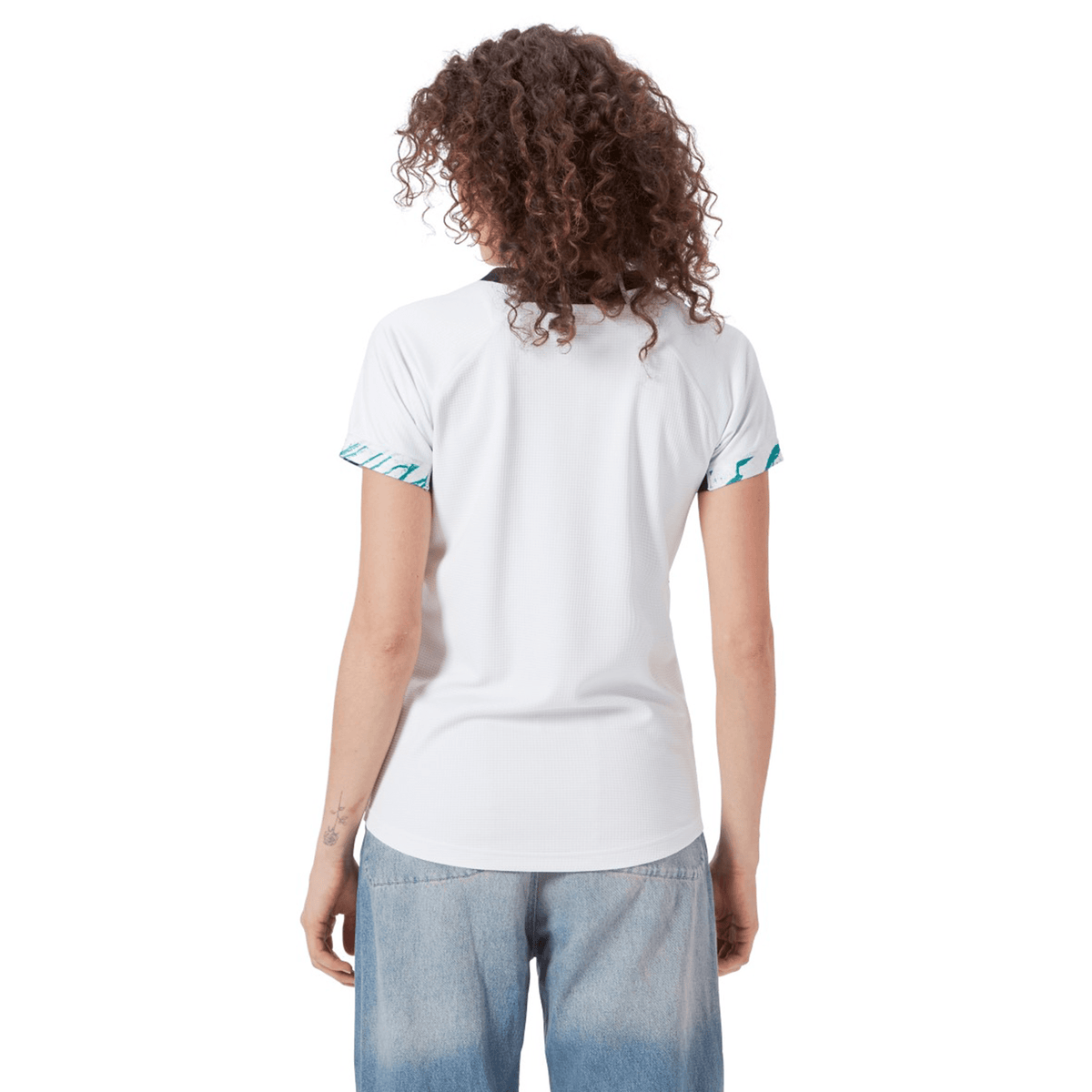 A person with curly hair faces away from the camera, wearing a white T-shirt with patterned sleeves, similar to the Women's Ireland 24 Alternate Pro Jersey by Canterbury, and blue jeans.