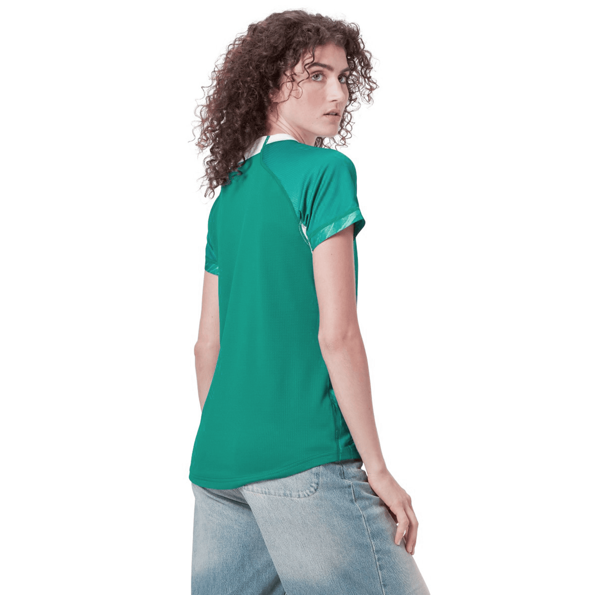 A person with curly hair wearing Canterbury&#39;s Women&#39;s Ireland 24 Home Pro Jersey in green and jeans stands facing away, looking over their shoulder.