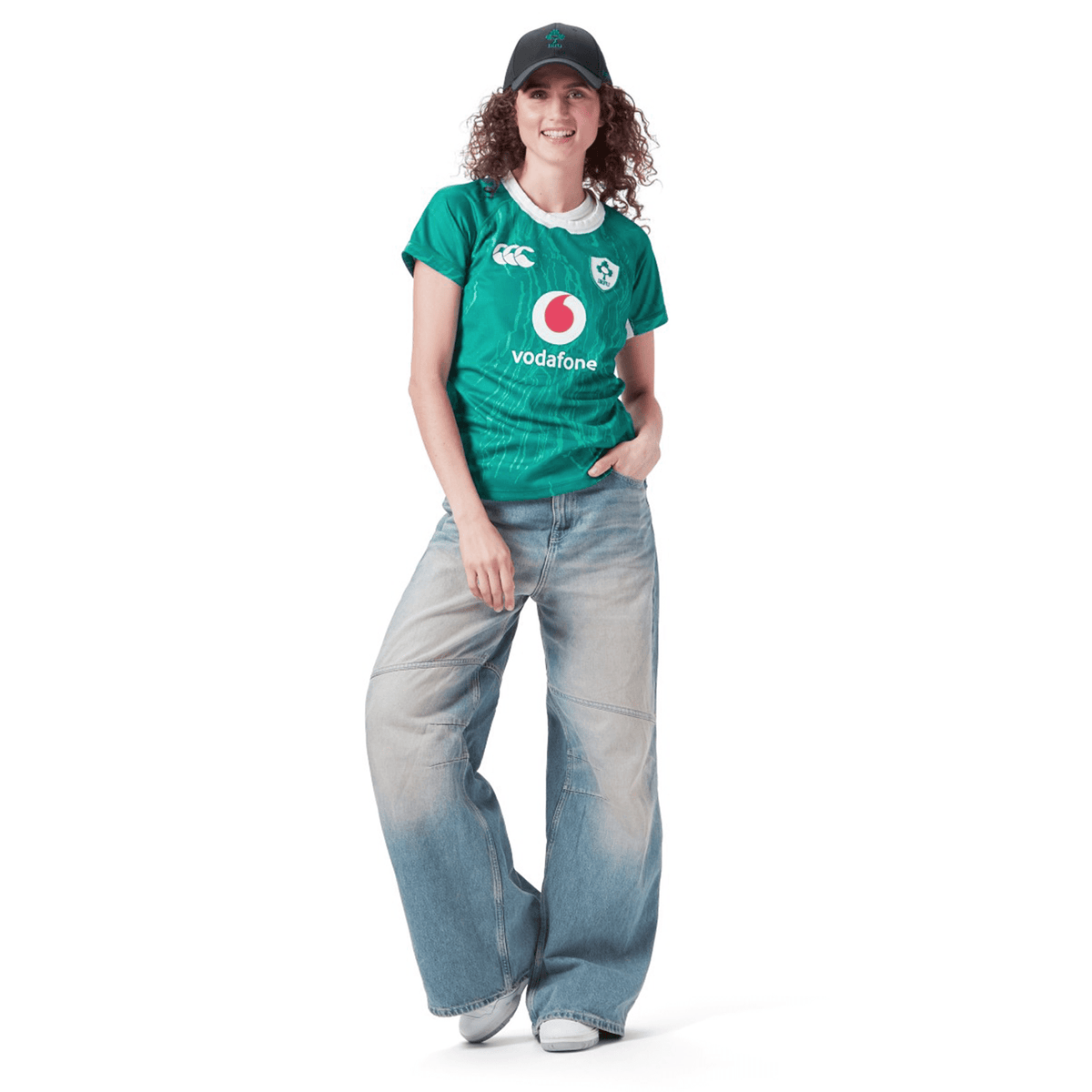Dressed in a Women&#39;s Ireland 24 Home Pro Jersey by Canterbury in vibrant green, blue wide-leg jeans, and a black cap, an individual stands confidently against a white background.