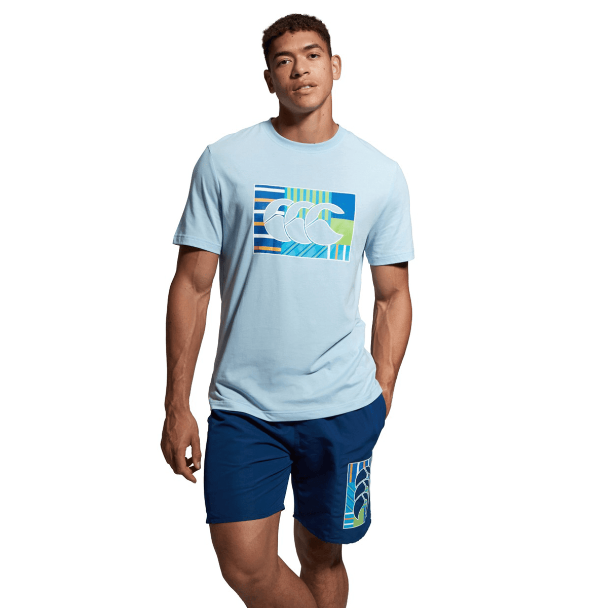 A person wearing a light blue Canterbury Canterbury Uglies T-Shirt with a graphic design and blue shorts stands against a white background.