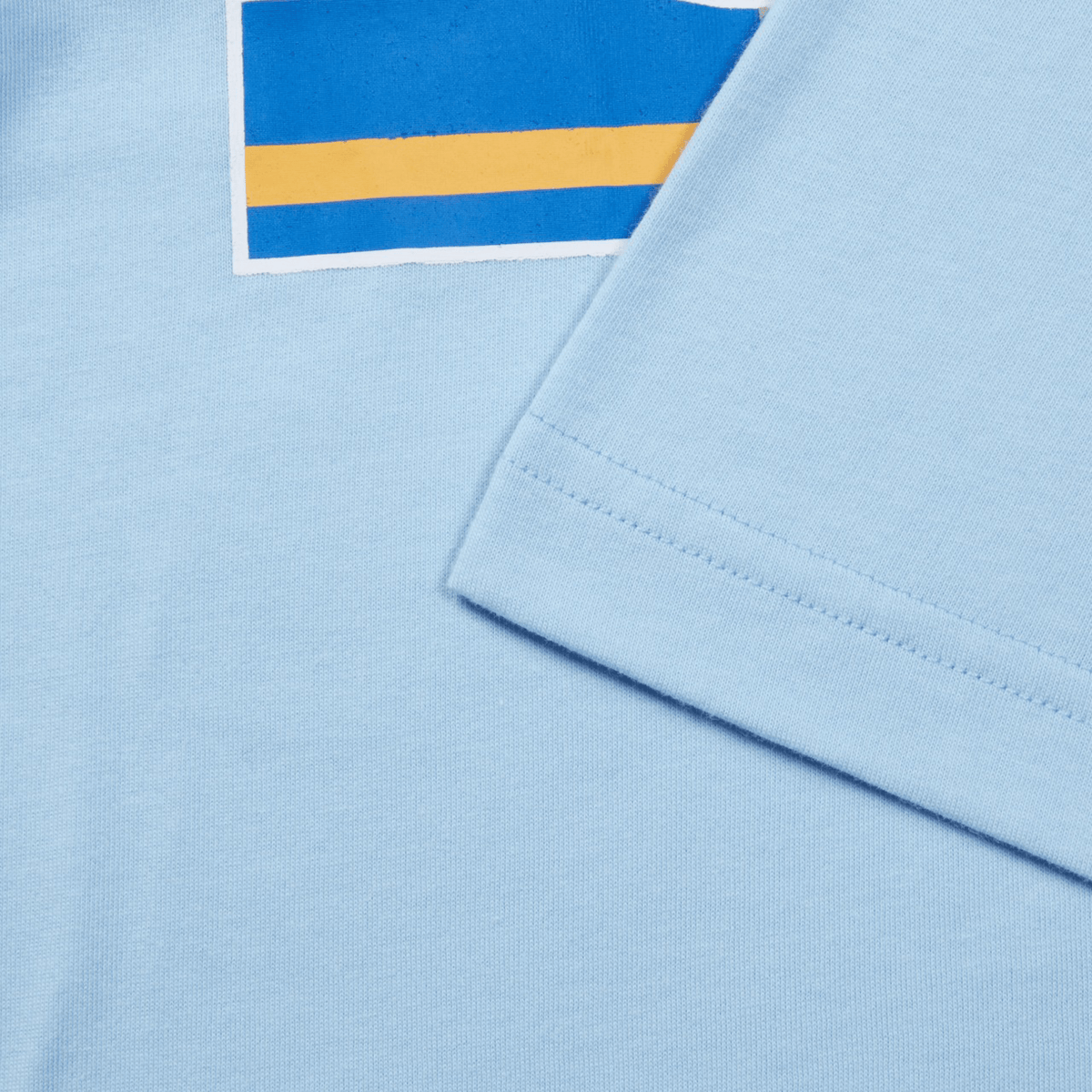 Close-up of a light blue Canterbury Uglies T-Shirt by Canterbury with a partial graphic print featuring blue and yellow stripes. The T-shirt's short sleeve is folded over.
