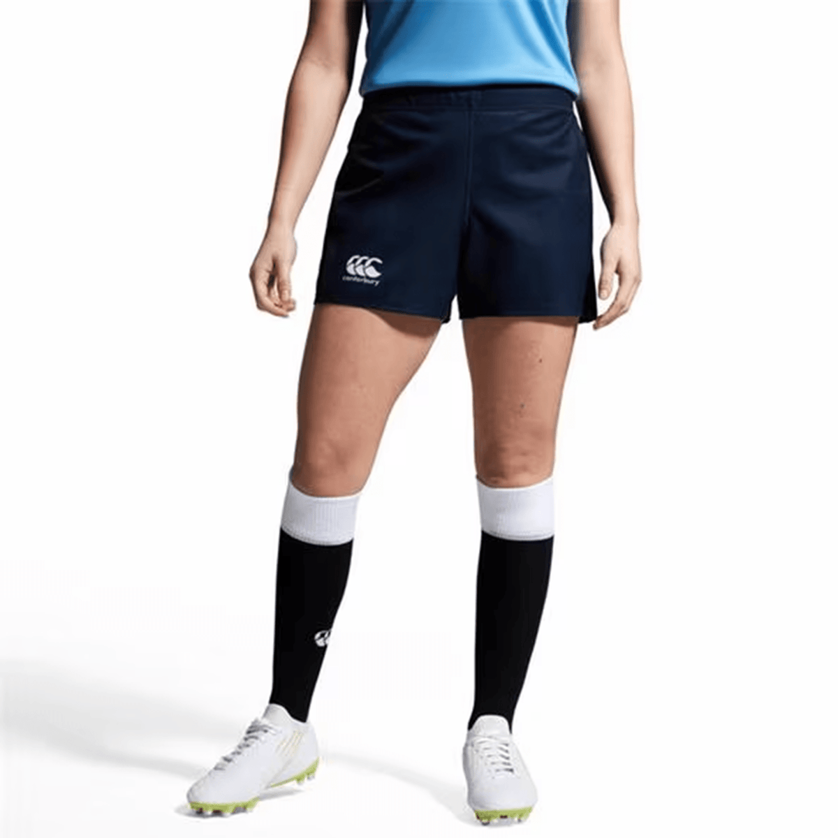 A person wearing a blue shirt with the Canterbury logo, Women&#39;s Canterbury Yokohama Shorts, white cleats, and black knee-high socks stands against a plain background.