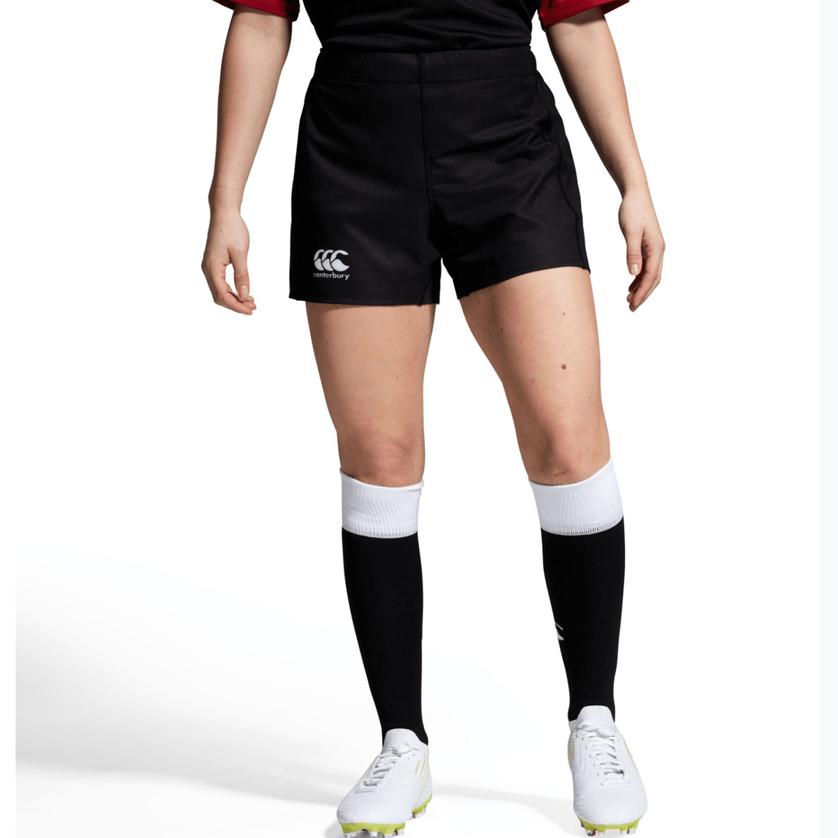 A person in a black Bundle Canterbury sports uniform with a white logo, long black socks with silicone grippers for support, and white cleats stands against a white background.