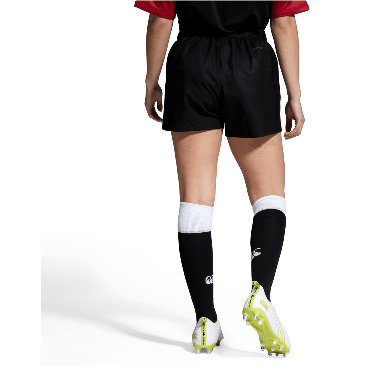An individual in a sports uniform stands on a white background, wearing Canterbury Yokohama shorts, a black shirt with red sleeves, knee-length black socks, and green cleats.