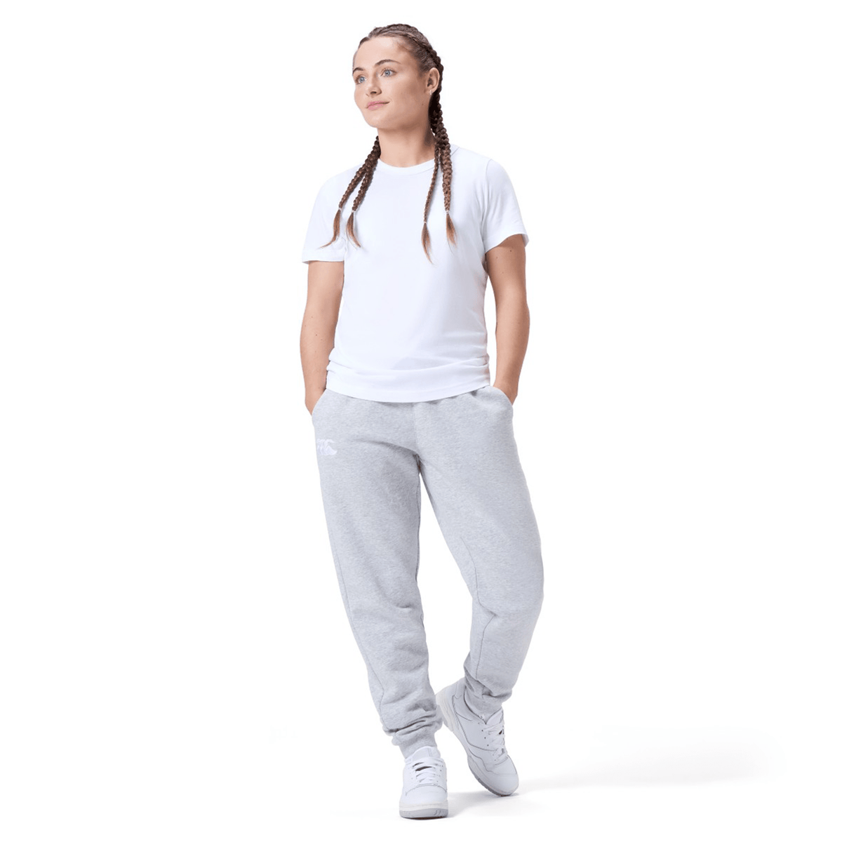 A person in a white t-shirt, Canterbury Women&#39;s Leisure Sweatpant by Canterbury, and white sneakers stands with hands in pockets, looking slightly to the side. Their hair is styled in braids, complementing the casual cotton blend fleece outfit perfectly.