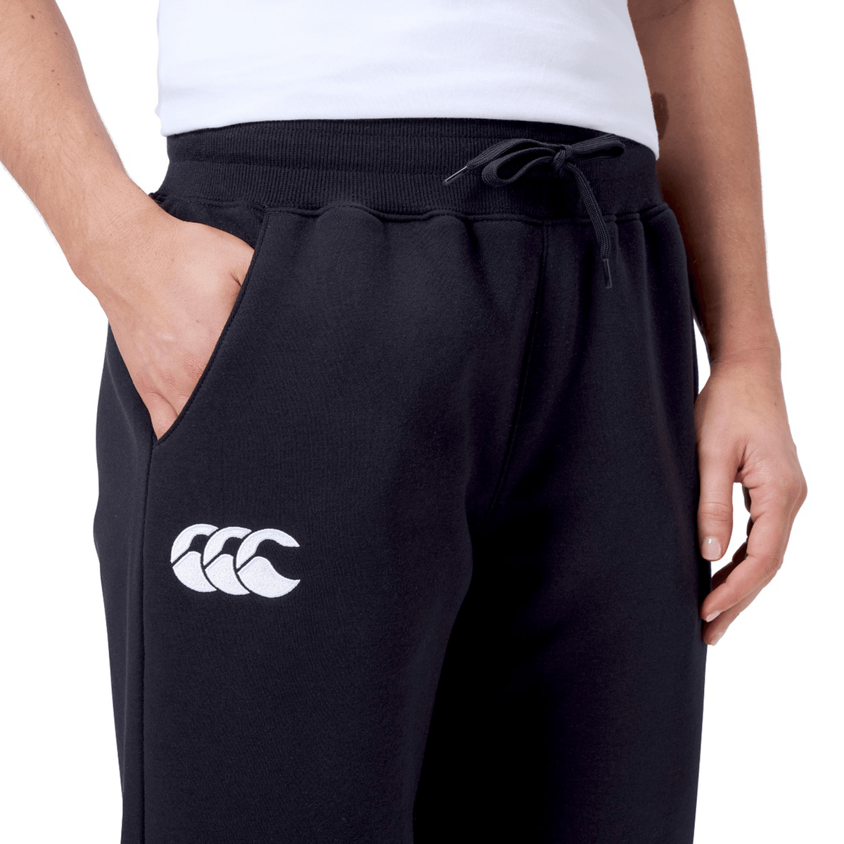 A person wears the Women&#39;s Leisure Sweatpant by Canterbury, made from a cozy cotton blend fleece in black, featuring a white Canterbury logo on the left thigh and has a hand in one pocket.