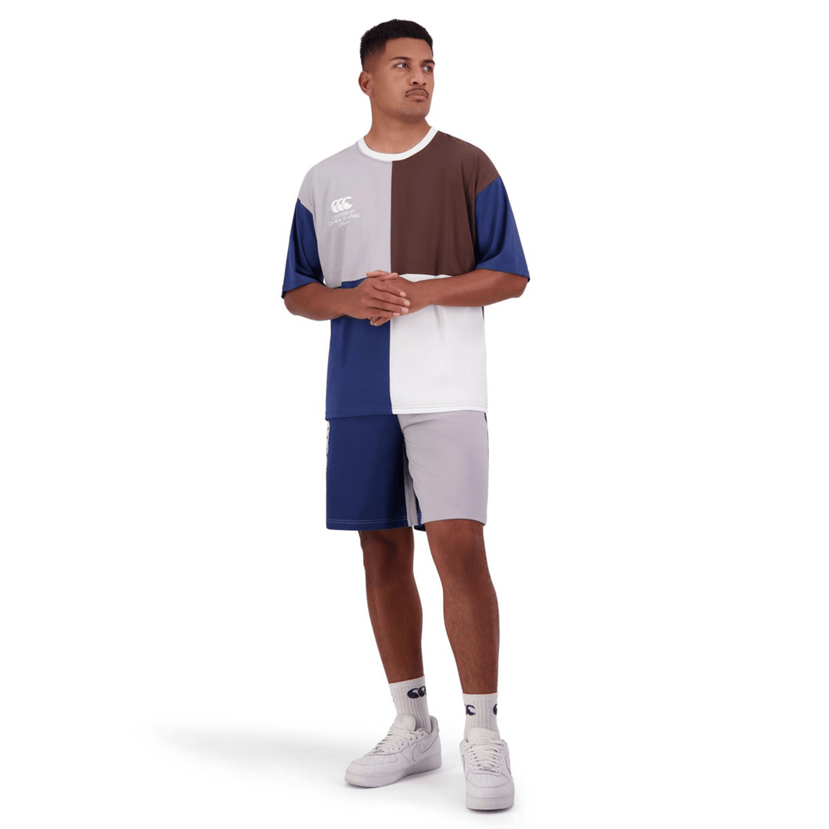 A person stands wearing a Canterbury Harlequin Block T-Shirt and matching shorts featuring an asymmetric design with shades of blue, brown, white, and grey. They are also wearing white shoes and socks. The background is plain white.