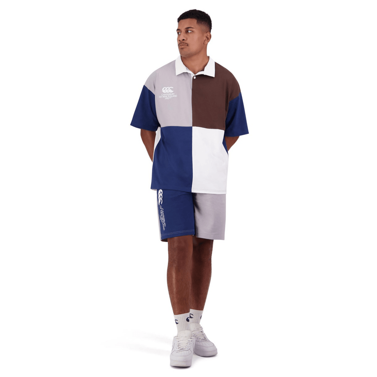 A man wearing a Canterbury Harlequin Block S/S Rugger polo shirt and shorts, standing with hands in pockets. The outfit features the distinctive color-block design in shades of blue, white, and brown. He completes the look with white sneakers and socks.