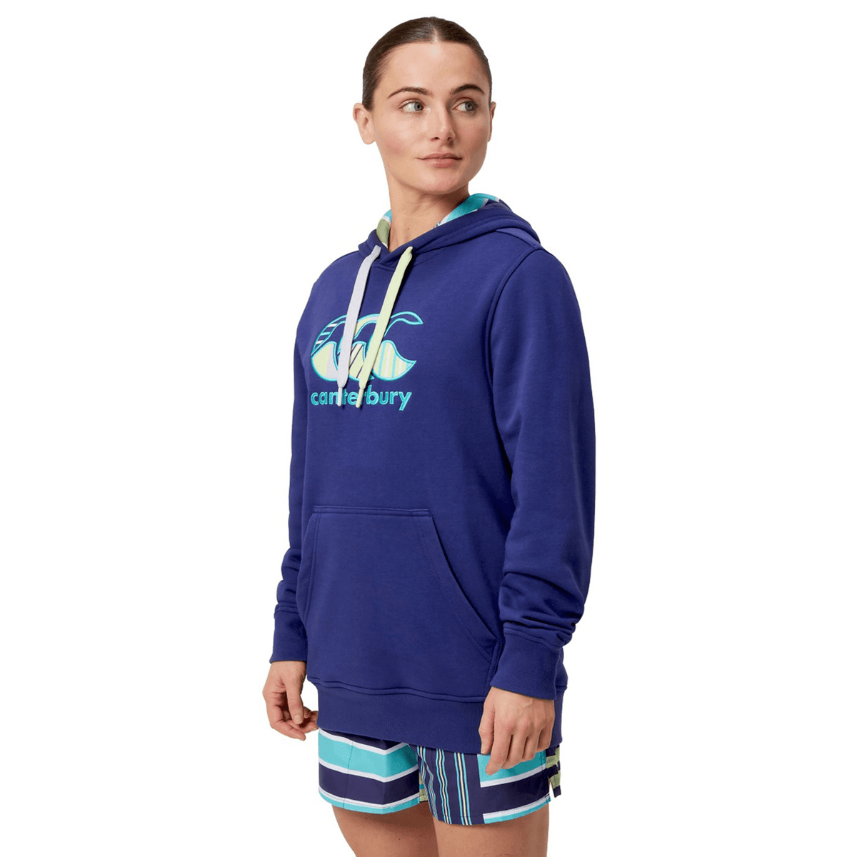 A person in a Canterbury Women&#39;s Uglies Hoodie with logo and striped shorts, standing against a white background, embodies Canterbury flair.