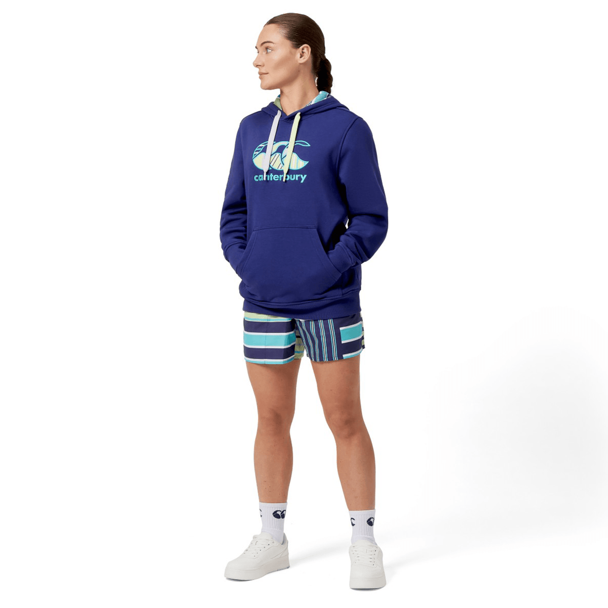 A person in a Canterbury Women&#39;s Uglies Hoodie in blue and striped shorts stands with hands in pockets, looking to the side. They&#39;re wearing white high-top sneakers and socks, exuding a laid-back vibe.