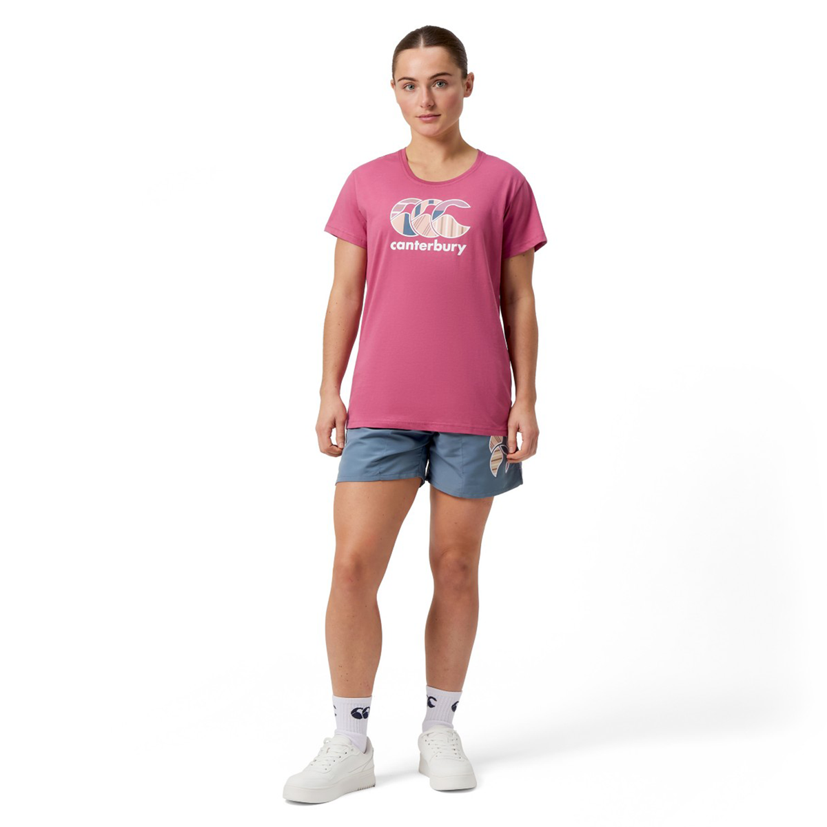 A woman wearing a pink Women&#39;s Canterbury Uglies T-Shirt by Canterbury, blue shorts, white socks, and sneakers stands against a plain white background, exuding a casual yet vibrant style reminiscent of Malaga Rose hues.