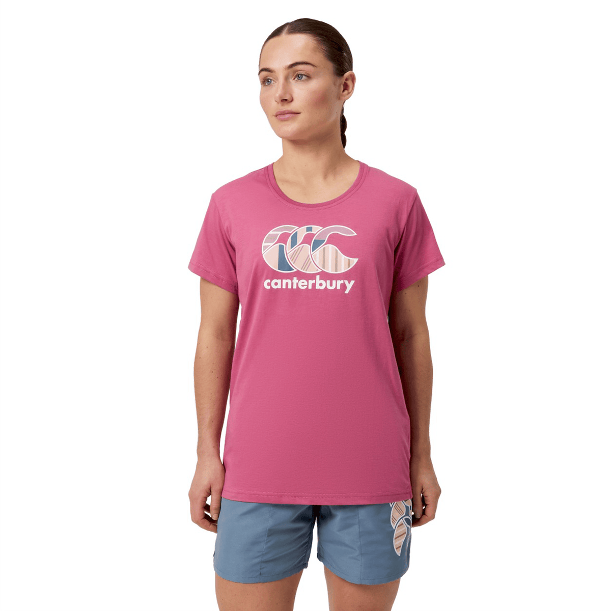 A person is standing against a white background, wearing a Women&#39;s Canterbury Uglies T-Shirt in pink and blue shorts.