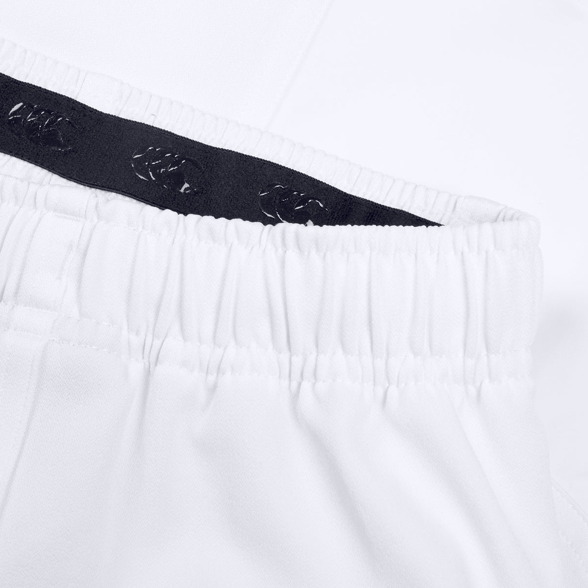 Close-up of a pair of white Canterbury Advantage Short 2.0 elastic-waist pants with a dark interior waistband lining featuring a repeating pattern. Made from recycled content, the fabric appears smooth and lightweight, reflecting Canterbury's commitment to performance sustainability.