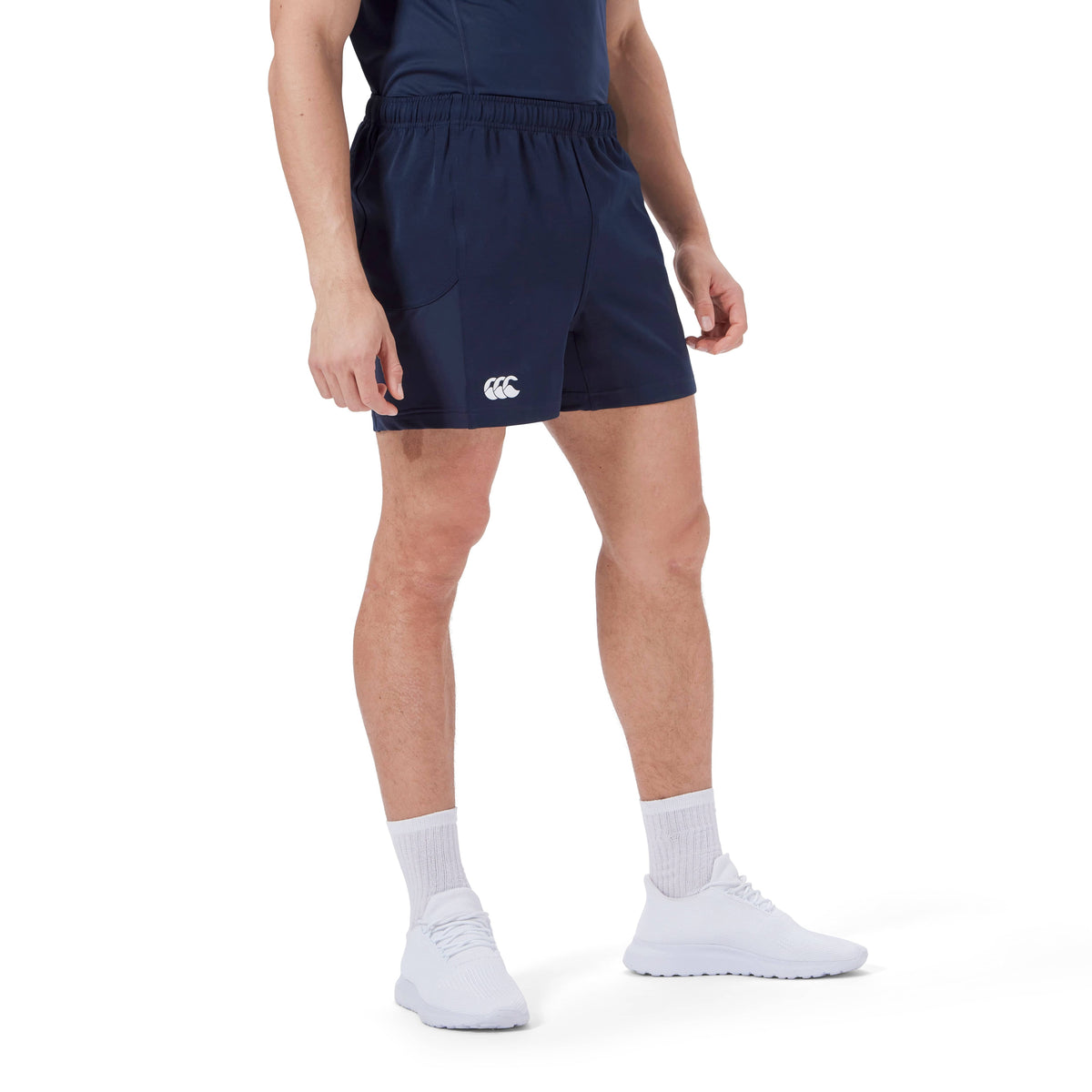 A person stands wearing a navy blue shirt and Canterbury Advantage Short 2.0 with white athletic shoes and socks, showcasing performance sustainability with recycled content.