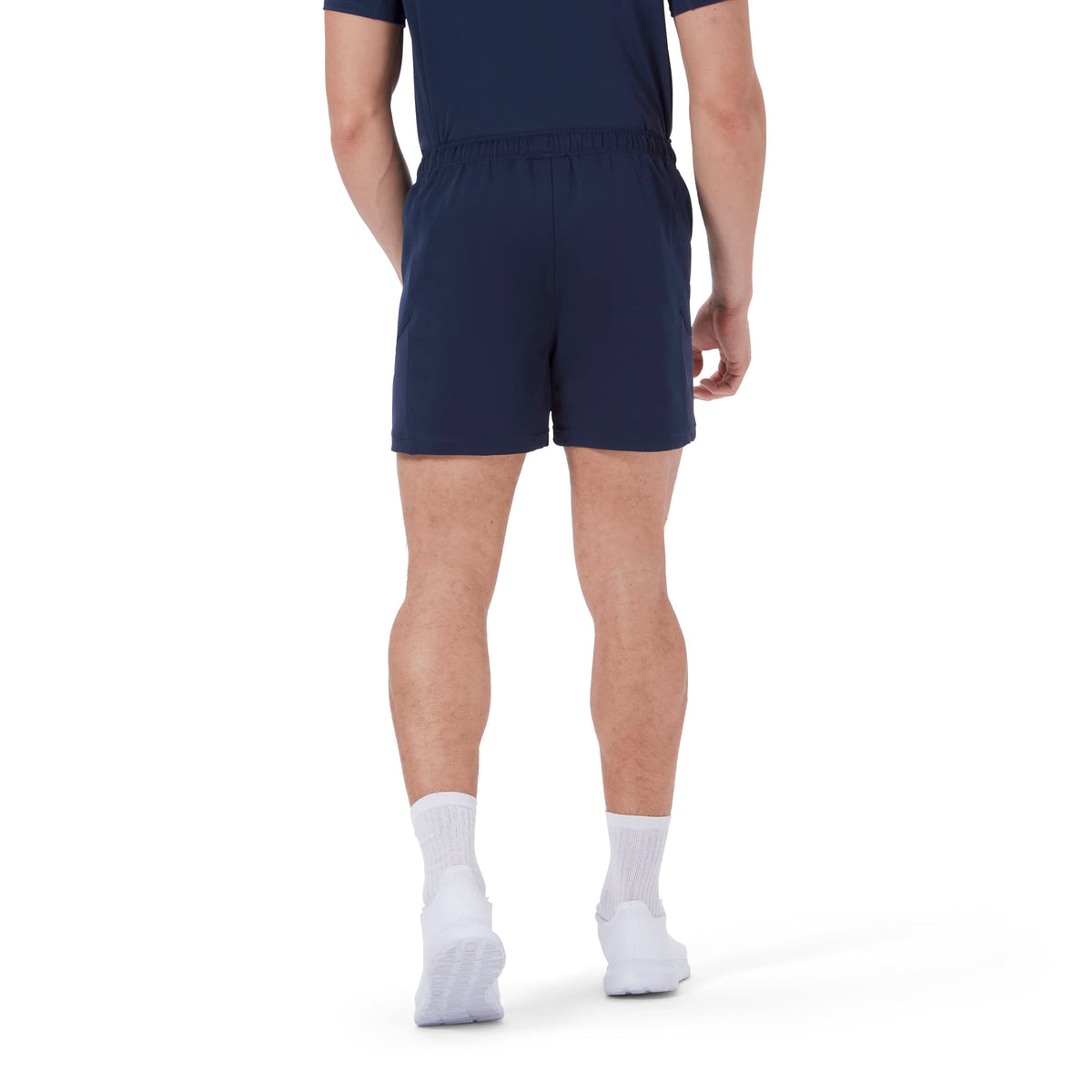 A person is standing with their back to the camera, wearing a navy blue Canterbury Advantage Short 2.0 outfit with matching shorts, white socks, and white shoes. The ensemble emphasizes both performance sustainability and style, incorporating recycled content for an eco-friendly choice.