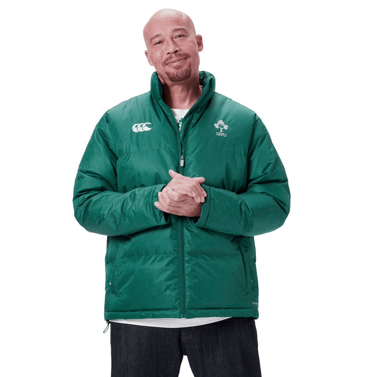 A bald man wearing a green Ireland rugby jacket and dark jeans stands with hands clasped, embodying the spirit of the Ireland 24 Puffer Jacket by Canterbury.