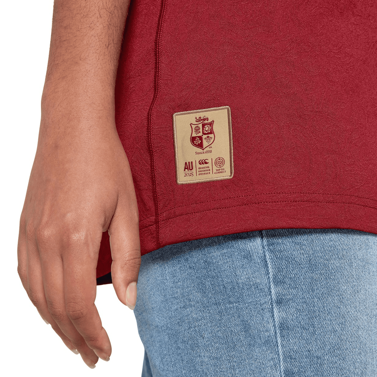 Close-up of a person wearing the British &amp; Irish Lions 2025 Players Jersey by Canterbury, paired with blue jeans. Only the lower torso and hand are visible.