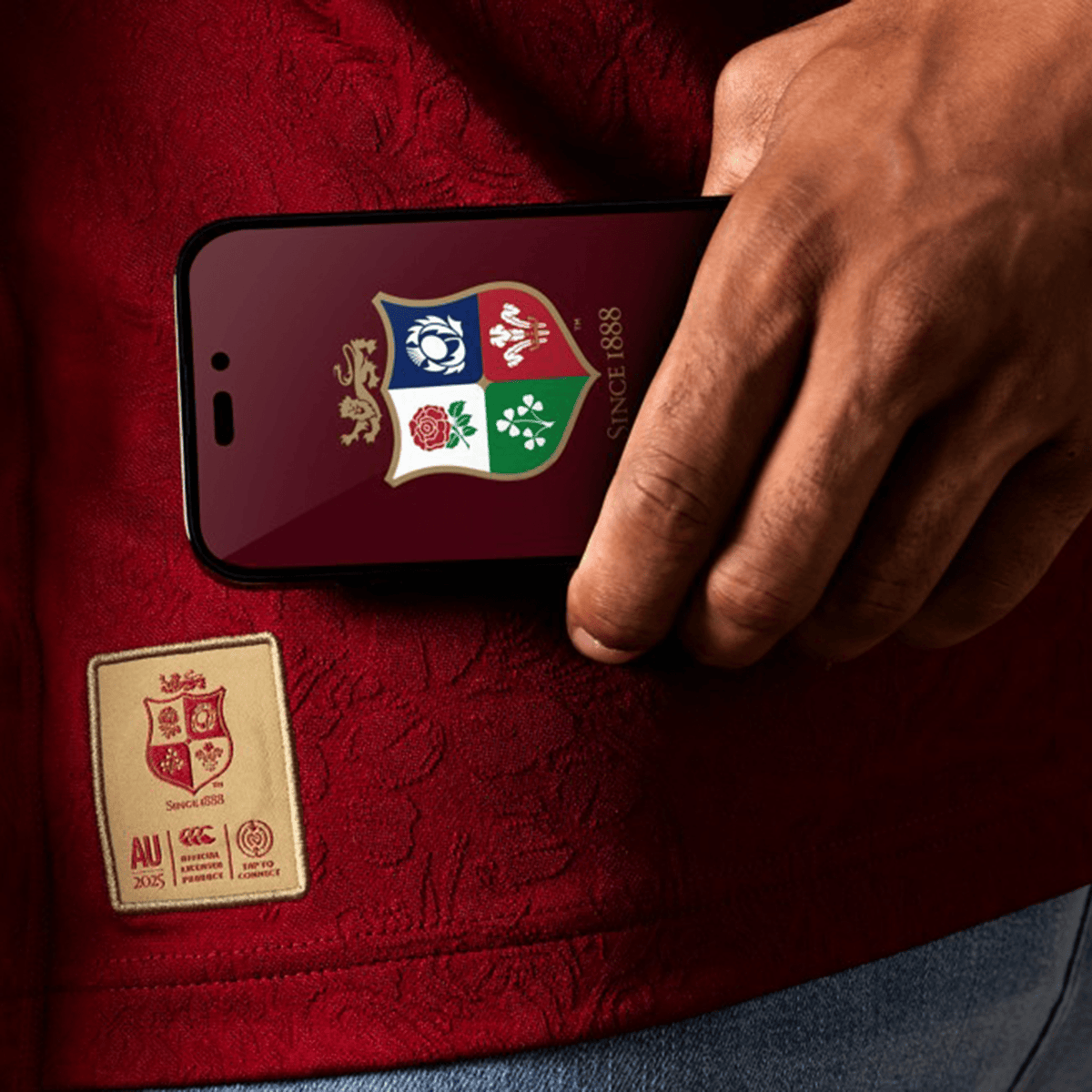 Close-up of a person holding a phone with a rugby-themed screen, wearing the red British &amp; Irish Lions 2025 Players Jersey by Canterbury, featuring a stitched emblem.