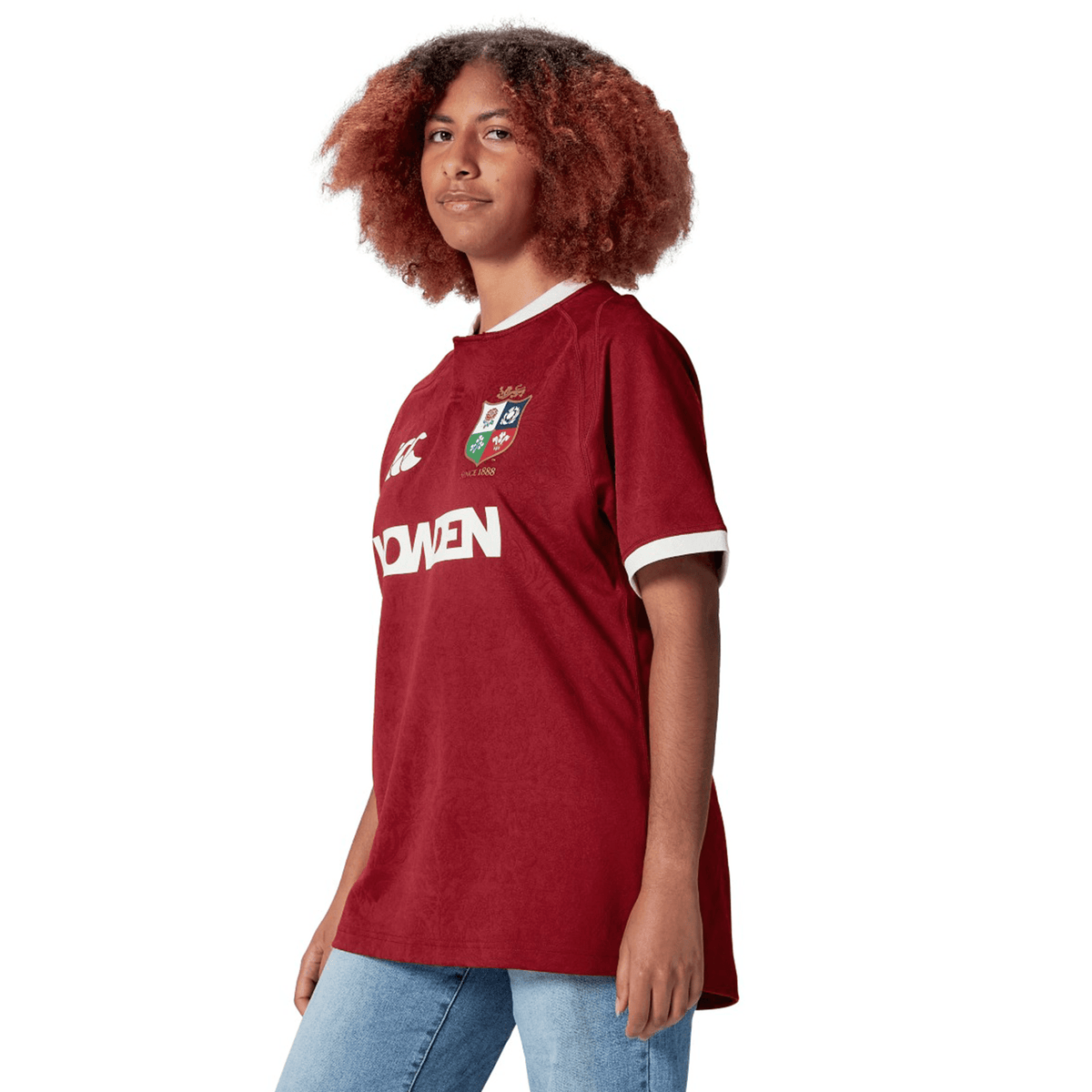 A person wearing a red British &amp; Irish Lions 2025 Players Jersey by Canterbury and blue jeans stands against a white background.