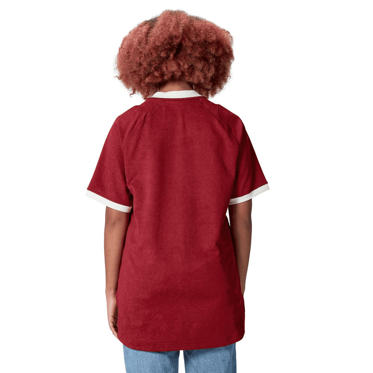 A person with curly hair, clad in a red British &amp; Irish Lions 2025 Players Jersey by Canterbury, featuring white cuffs, is seen from behind.