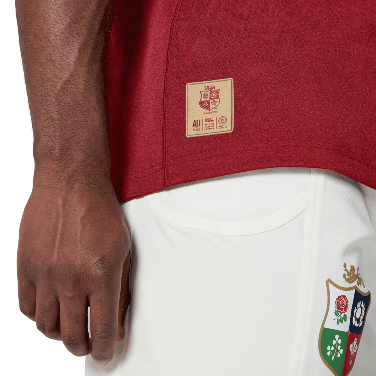 Close-up of a person wearing the British &amp; Irish Lions 2025 Players Jersey by Canterbury, a red sports shirt crafted from moisture-wicking fabric featuring a gold label, paired with white shorts adorned with a colorful emblem.