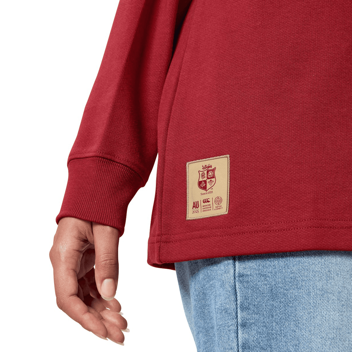 A person is dressed in the British &amp; Irish Lions 2025 Long Sleeve Classic Jersey by Canterbury, featuring a logo and patch near the hem. They have their left hand visible and are wearing light blue jeans. This jersey is designed with moisture-wicking fabric for added comfort.